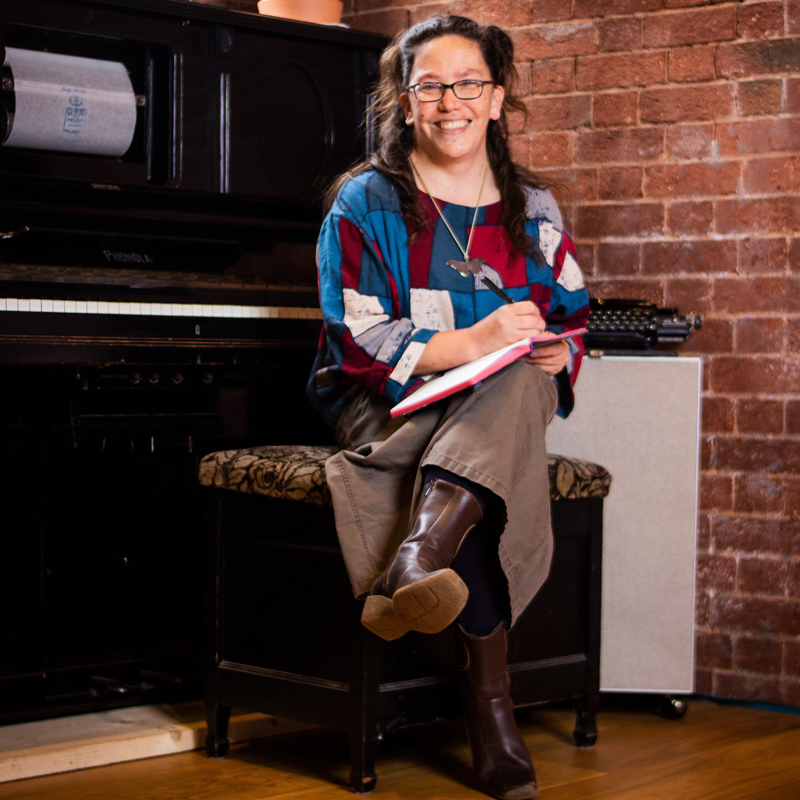 Poet harula ladd sits on a piano stool, smiling at the camera holding a notebook.