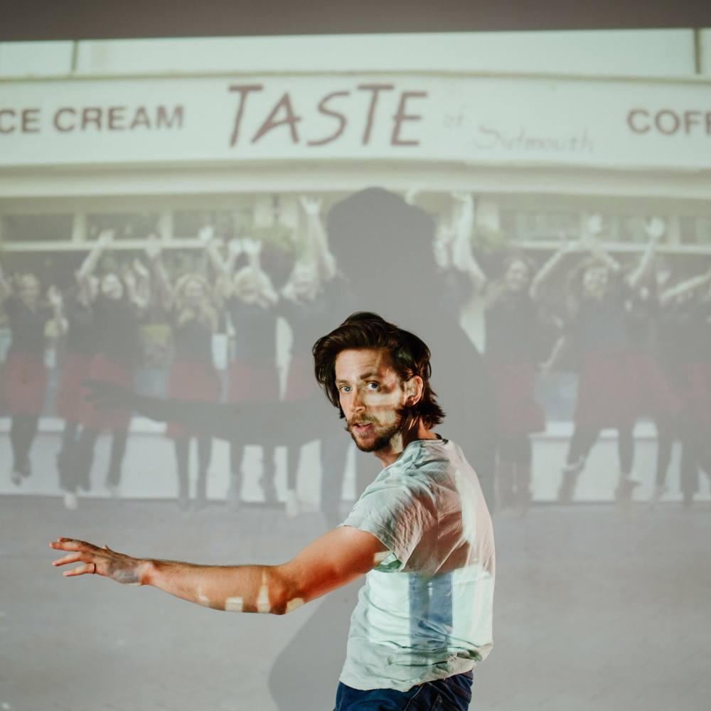 Performer Lewys Holt stands in front of a projector, wearing a white tshirt and looking at the camera.